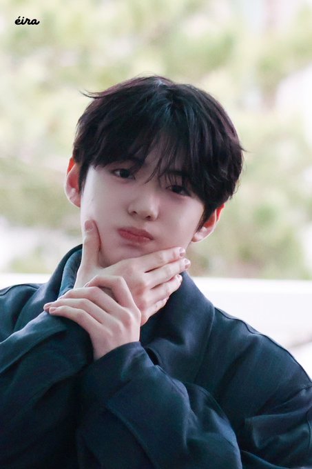 A young man with dark hair poses thoughtfully with his hands under his chin, wearing a dark jacket against a softly blurred outdoor background. The watermark "éina" appears in the upper left corner, and the image accompanies a post tagging #정상현 (Jung Sanghyeon) and #ALPHADRIVEONE (#ALD1), suggesting this is a fan-taken photo of the Alpha Drive One member.