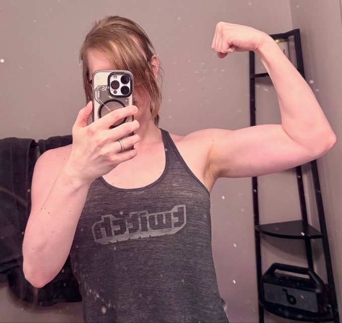 A person wearing a dark gray 'DBZ' tank top takes a mirror selfie while flexing their bicep, holding a smartphone that partially covers their face.