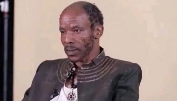 A Black man with a short haircut and stubble wearing a brown leather jacket with decorative stitching, photographed against a plain beige wall.