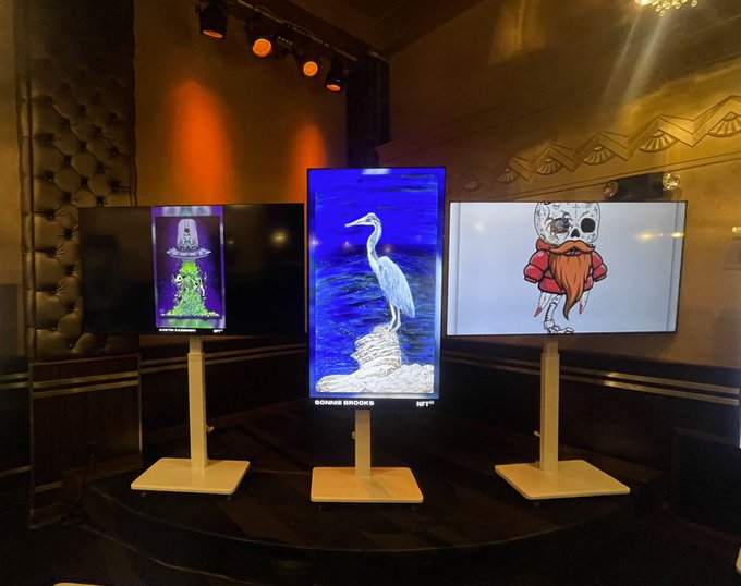 Three digital screens stand on pedestals in a dimly lit gallery, displaying digital art including a green creature, a heron ('SOANRS BROOKS' NFT), and a skull-headed character in a red hoodie.