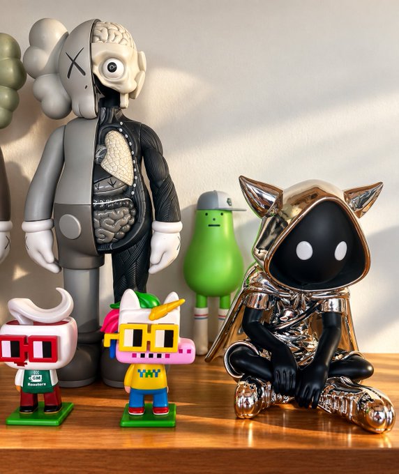 A display of designer toys and art figures on a wooden surface, including a grey dissected figure, a gold-hooded character, and several smaller whimsical figures.