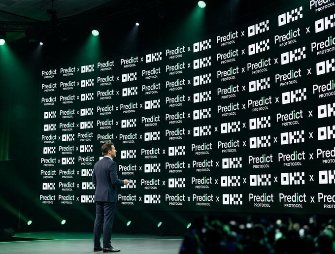 A man in a suit stands on stage in front of a large screen displaying 'Predict x OKX PROTOCOL' branding at a corporate event.