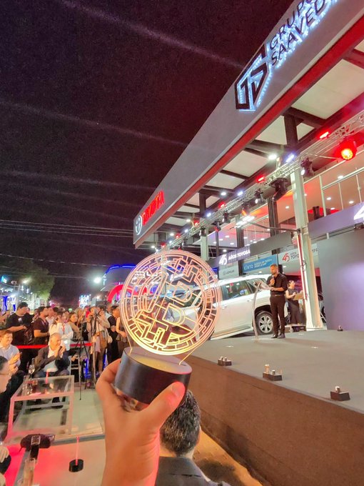 A hand holds an illuminated trophy at what appears to be a tech event, with a building labeled 'SAVED' and a Toyota booth visible in the background.