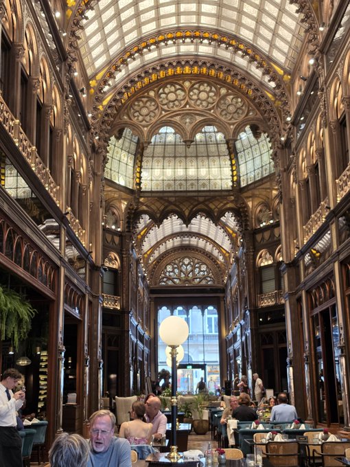 The ornate, cathedral-like interior of a grand building featuring intricate ironwork, arched stained-glass ceilings, and chandeliers, with patrons dining below.