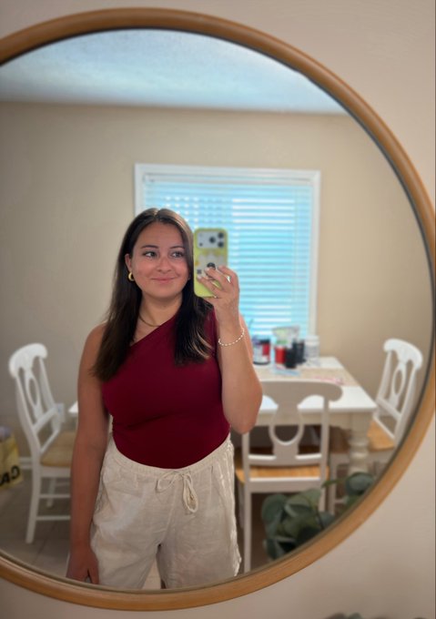 A young woman takes a mirror selfie in a round wooden-framed mirror, wearing a burgundy one-shoulder top and cream-colored drawstring pants, smiling softly while holding a phone with a light-colored case. The "gm too everyone who gms" caption suggests a cheerful morning greeting to fellow early risers, matching her warm, relaxed pose in what appears to be a sunlit dining area.