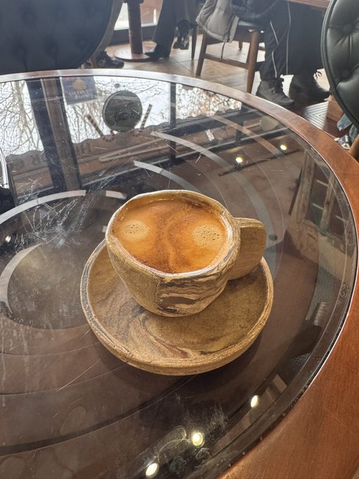 A rustic ceramic cup and saucer holding a freshly pulled espresso sits on a glass-topped wooden table in what appears to be a café setting.