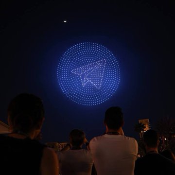 A nighttime scene showing a crowd of people watching a drone light formation creating a paper airplane/paper plane shape glowing in blue against the dark sky.