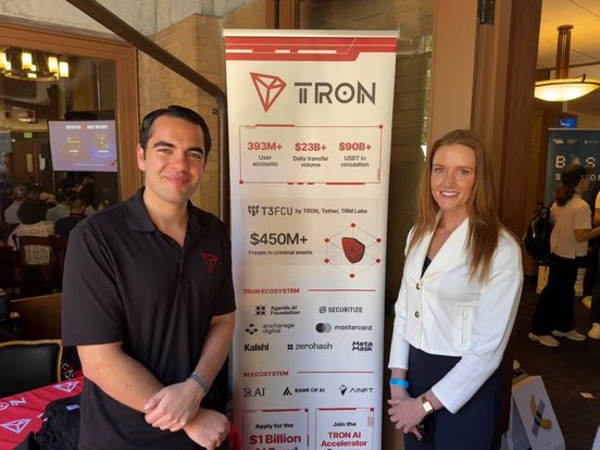 Two individuals stand next to a TRON banner displaying its ecosystem statistics and initiatives, such as the TRON AI Accelerator, at the Blockchain Applications Stanford Summit (BASS).