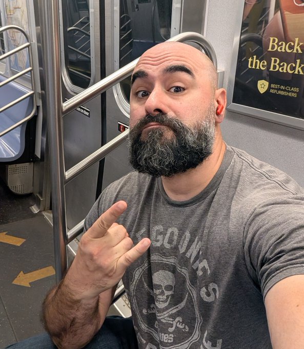 A bald man with a gray-flecked beard takes a selfie on a New York City subway, pointing at himself while wearing a Guns N' Roses skull t-shirt. Behind him, an advertisement promotes "Back the Back" and "Best-in-Class Refurbishers," capturing his travels to NYC for NFTNYC.