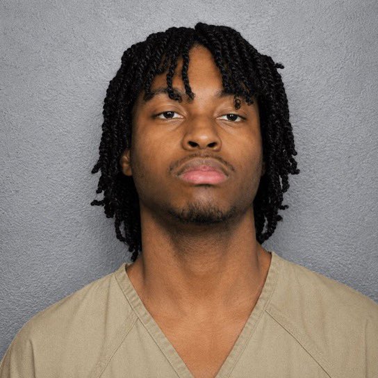 A mugshot of a young Black man with long twisted locs and a neutral expression, wearing a beige V-neck garment against a gray background—appearing to be the individual accused of stealing six figures from a cancer patient through a malware-infected Steam game that compromised the victim's passwords and private keys.