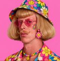 A stylized portrait of a person wearing a blonde bob wig, pink round sunglasses, a colorful floral hat, and matching floral clothing against a bright pink background.