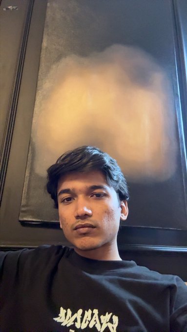 A young man in a black t-shirt poses for a selfie, with a blurred painting visible on the wall behind him.