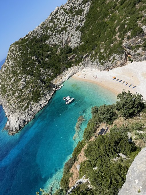 A stunning aerial view of a secluded turquoise cove beach nestled between dramatic rocky cliffs in Albania, with two small boats anchored in the clear waters and sun loungers arranged along the sandy shore. The post connects this serene, recharging escape from Albania to the creator's renewed focus on sending $TIBBIR to the top 10, pairing their refreshing break with a call to action to push the token upward.
