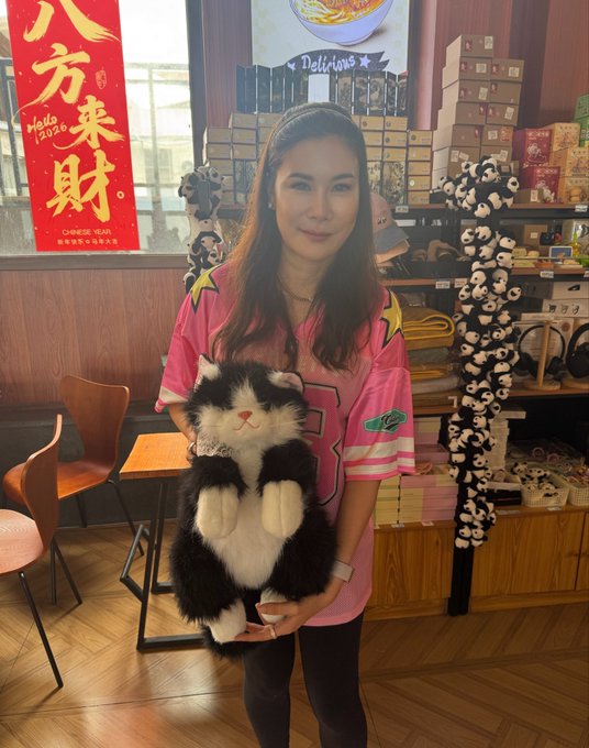 A woman wearing a pink shirt and headband holding a black and white stuffed cat in a shop with Chinese New Year decorations (八万来财) and numerous plush toys visible.