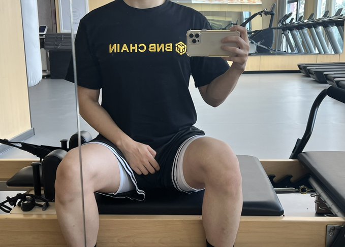 A person sitting on a Pilates reformer machine wearing a black BNB Chain t-shirt (backwards text reading 'NAHC BNB') and black shorts with white trim, taking a mirror selfie with an iPhone.