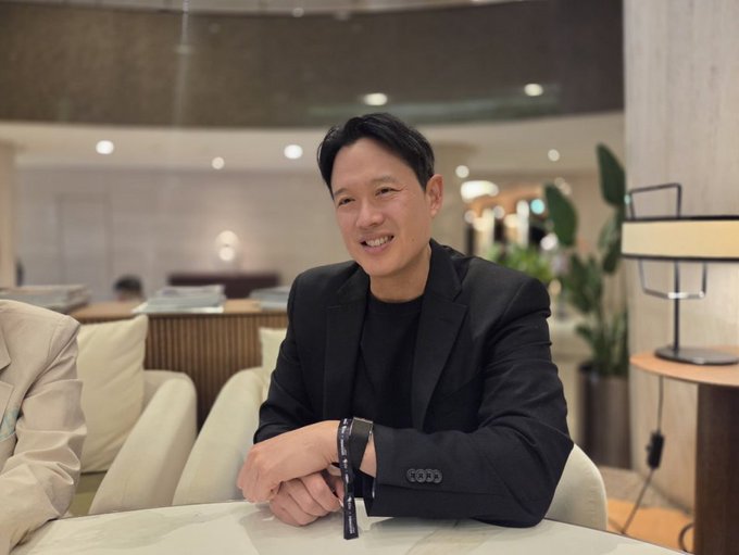 An East Asian man in a black blazer and black t-shirt smiles warmly while seated on a light-colored sofa, wearing a wristband and holding sunglasses in a modern lounge or office setting.