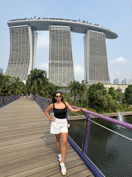A woman poses on a wooden bridge with the iconic Marina Bay Sands hotel towering in the background. The image captures the Singapore vibes referenced in the "still in Singapore" post, showcasing one of the city's most recognizable landmarks.