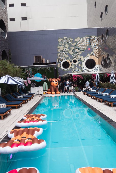 A vibrant poolside scene features inflatable popsicle floats drifting in turquoise water, with a large teddy bear statue and bold cartoon-style mural as the backdrop, perfectly capturing the playful, weekend-ready spirit of "Happy Friday" and "GM" energy.