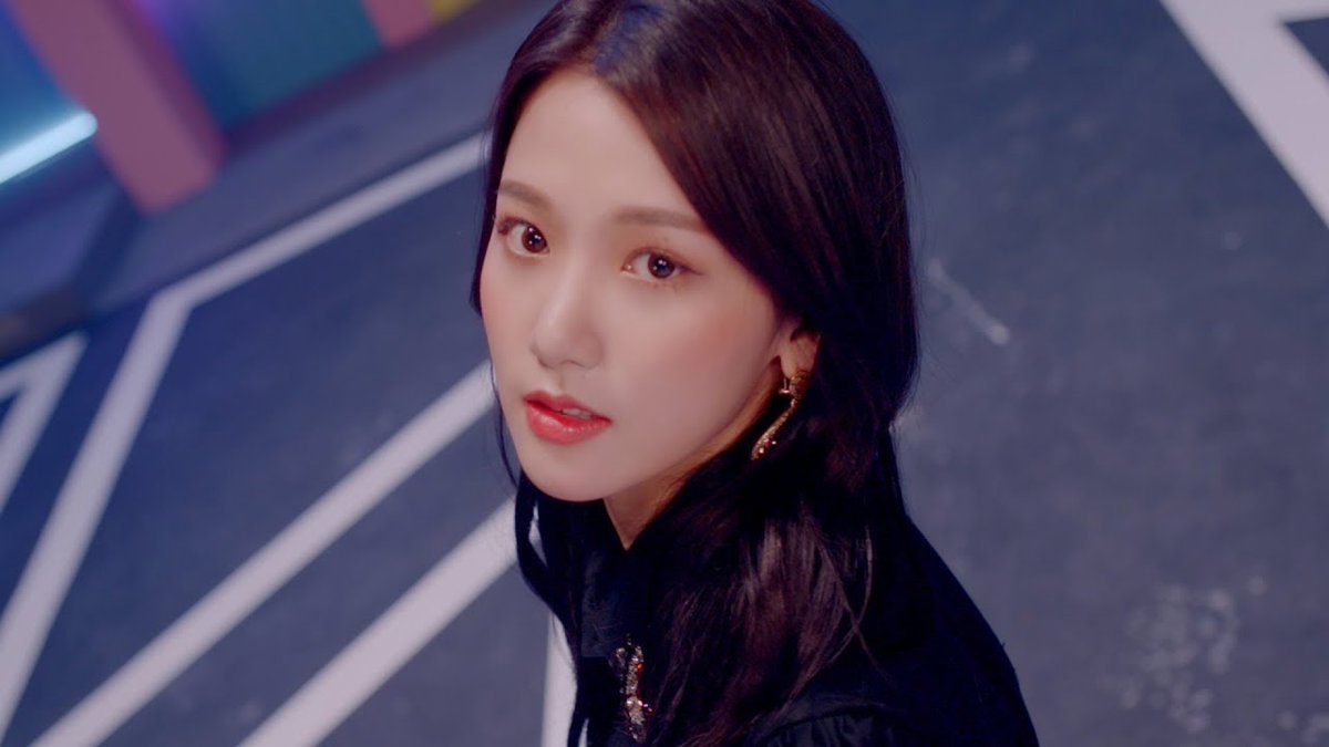 A portrait of a young woman with long dark hair, wearing red lipstick and dangling earrings, looking toward the camera against a blue floor background.