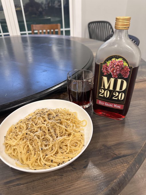 The image shows a plate of spaghetti with cheese, accompanied by a glass of wine. The wine bottle has "MD 2020" written on it, indicating that it's a red wine from MD. There are two chairs around the dining table where the meal is being served. One chair is located near the left side of the table and another one is positioned closer to the right side. A bowl containing spaghetti can be seen in front of the wine bottle, suggesting that someone has already started eating their meal.