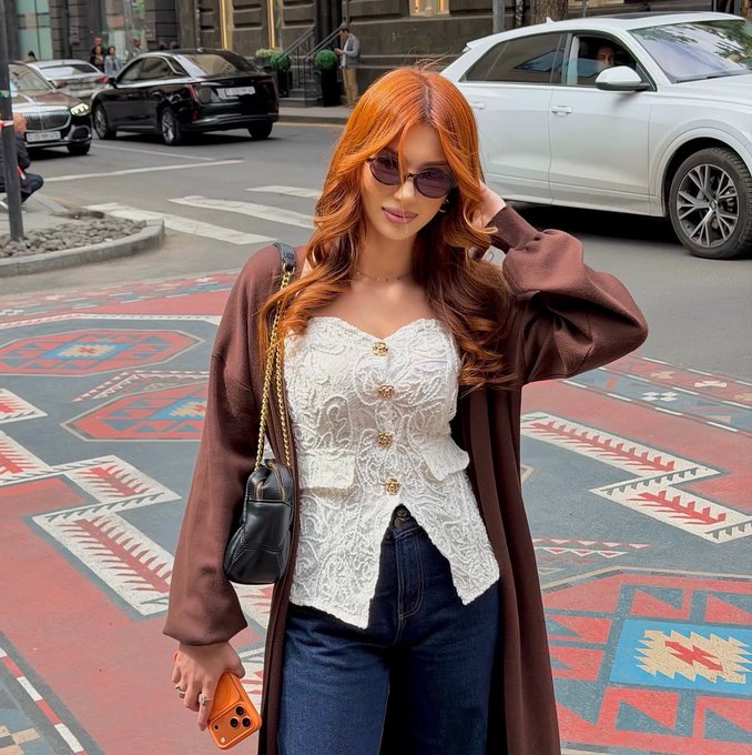 A young woman with long red hair and sunglasses stands on a city street wearing a white lace top, brown cardigan, and jeans, while holding an orange smartphone and a black purse.