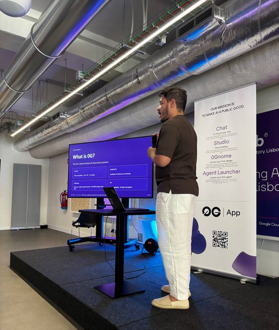 A man in a brown t-shirt and white pants presents on stage next to a screen displaying 'What is DG?' alongside a banner for 'OG App' that says 'OUR MISSION IS TO MAKE AI A PUBLIC GOOD'.