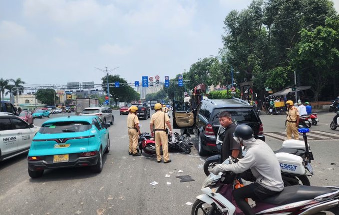 A street scene in Vietnam showing traffic police in yellow uniforms attending to a motorbike accident, with a bright turquoise VinFast taxi and various other vehicles on a busy road.