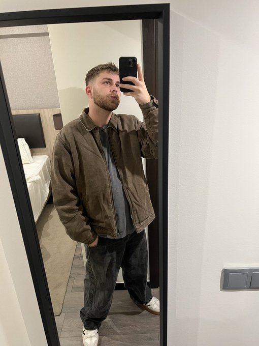 A man with a beard takes a mirror selfie in a hotel room, wearing a brown suede jacket, gray shirt, and dark jeans while holding up a black smartphone. The casual, polished outfit and morning-setting suggest he's ready to greet his crypto audience and collect $SOL wallet addresses for a tip drop.