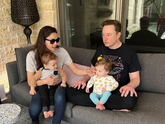 A family portrait showing Elon Musk sitting on a gray couch with a woman wearing sunglasses, both holding young babies on their laps in what appears to be a living room.