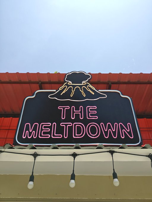 A neon sign reading 'THE MELTDOWN' featuring an illustration of an erupting volcano, mounted on a building under a red tiled roof.