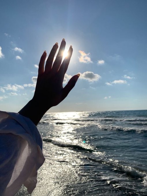 A silhouetted hand reaches toward the bright sun over a sparkling ocean, with a white sleeve visible against the blue sky and waves.