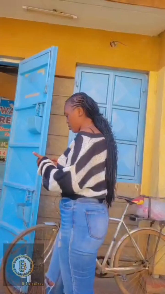 The image shows a woman standing next to a bicycle, wearing a black and white striped sweater. She has long hair and is holding a cell phone in her hand. The background features a yellow building with blue doors. There are also two bicycles visible in the scene - one near the woman's feet and another further away from her.