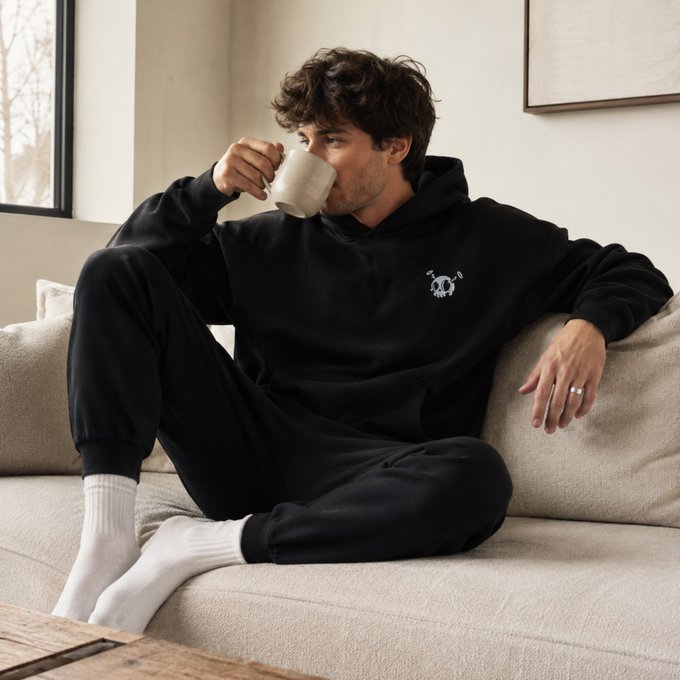 A young man with curly hair relaxing on a beige couch, drinking from a white mug while wearing a matching black hooded sweatshirt and sweatpants with white socks.