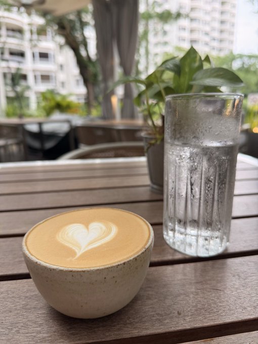 A latte with heart-shaped foam art sits on a wooden table next to a glass of water, with potted plants and buildings in the background.