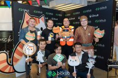 The image features a group of people posing for a photo behind a black backdrop adorned with a cartoon cat. The group consists of seven individuals, each holding up a sign or a small object that says "bitkub" or "bitcoin". They are all smiling and appear to be enjoying themselves at the event. In the background, there is a banner displaying the words "Zircuit", which suggests that this gathering might be related to Bitcoin or cryptocurrency-related activities. The overall atmosphere seems to be one of fun and excitement as people come together for an engaging and entertaining experience.