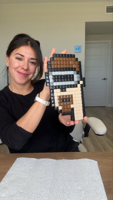 A smiling woman holds up a pixel art creation made of small colored blocks that resembles a character with brown hair and armor.