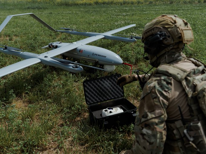 A soldier in camouflage gear and helmet operates a white fixed-wing 'PEREGRIN' drone and its controller in a foam-lined case on a grassy field.