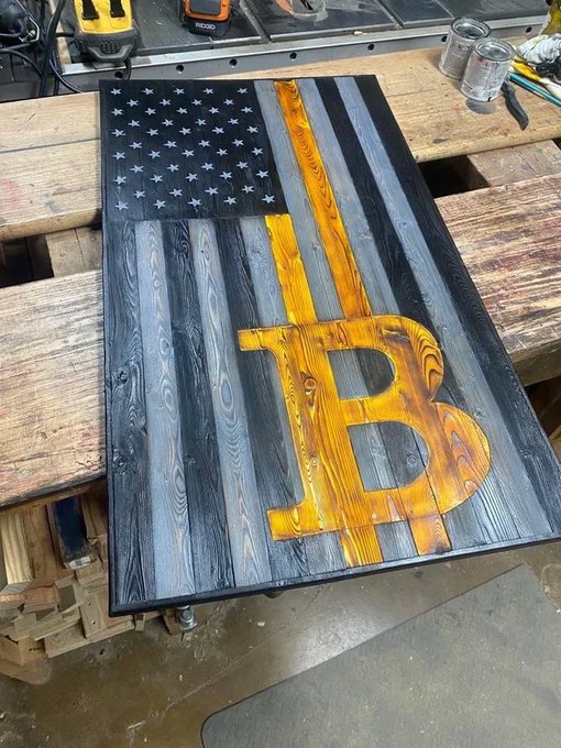 A wooden American flag features a Bitcoin symbol prominently displayed in gold where the stripes would be. Combining the patriotic imagery with cryptocurrency symbolism visually reinforces the message that "Bitcoin is freedom," suggesting digital currency as a tool of financial independence and liberty.