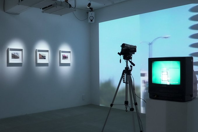 An art gallery installation featuring three framed photographs on a wall, a video camera on a tripod, and a television displaying a green-tinted video.