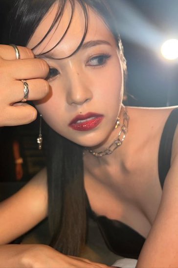 A close-up portrait of a woman with red lipstick, wearing a black top and silver jewelry, with her hand resting near her forehead.