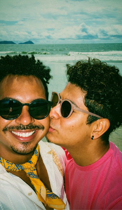 A vintage-style photograph capturing a joyful moment on a beach, showing a smiling man with sunglasses while another person in a pink shirt affectionately kisses his cheek.