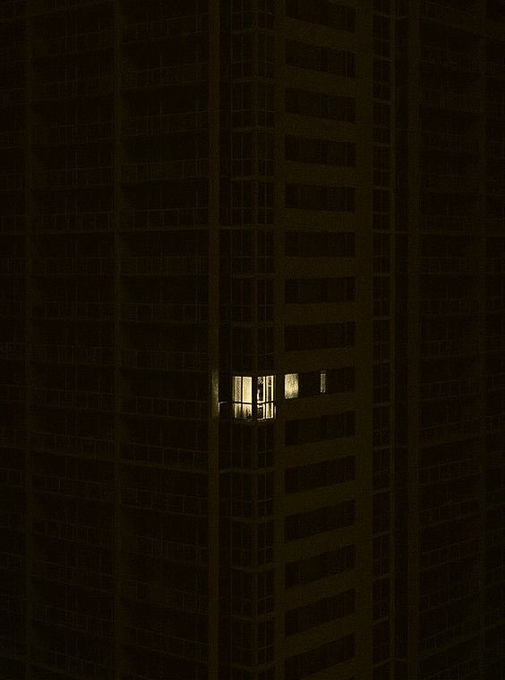 A nighttime photograph of a tall apartment building with a single illuminated window on an otherwise dark floor.