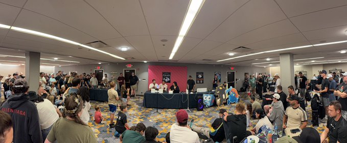 A panoramic view of a crowded convention room with people sitting on a patterned carpet watching a presentation at tables in front of a pink backdrop.