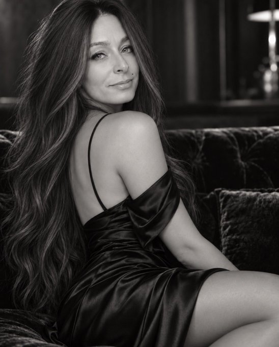 A black-and-white portrait of a woman with long wavy hair wearing a dark satin off-the-shoulder dress, seated on a tufted velvet sofa.