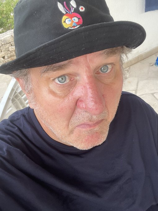 A close-up selfie shows an older man with blue eyes wearing a black t-shirt and a black bucket hat featuring a colorful cartoon design.