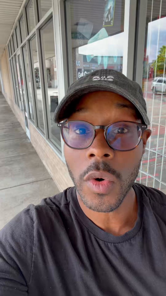 The image features a man wearing glasses, a black t-shirt, and a baseball cap with the word "Topps" written on it. He appears surprised or shocked as he takes a selfie using his phone's camera. The background of the photo shows a building, suggesting that they are taking the picture in an urban setting. The post includes text such as "Veefriends Topps chrome Day!!! @VeeFriendsCards @veefriends".