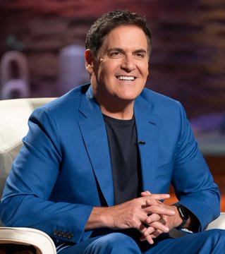 Mark Cuban smiling while wearing a blue blazer over a black shirt, sitting on a white chair in what appears to be a TV studio setting.