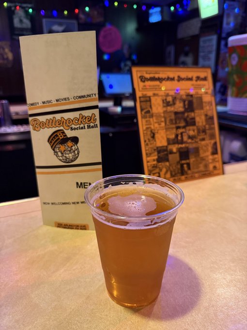 A plastic cup of beer sits on a table in front of a menu for 'Bottlerocket Social Hall' with a cartoon rocket logo, in a venue with string lights.