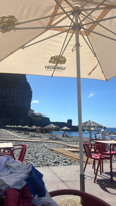 A beachside view from under a "Sombras del Sol" umbrella overlooking a rocky shoreline with palm-thatched parasols and red chairs, while the post "Gm fam Drop SOL addy to win" ties in with the umbrella branding "Sombras del Sol" as a play on the Solana (SOL) crypto address request.