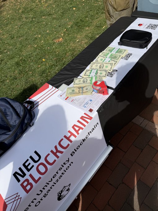 A table draped with a "NEU Blockchain - Northeastern University Blockchain Organization" banner displays stacks of dollar bills, informational flyers, and a tape measure, set up as a recruitment or fundraising booth likely at a campus event encouraging students to join the organization.